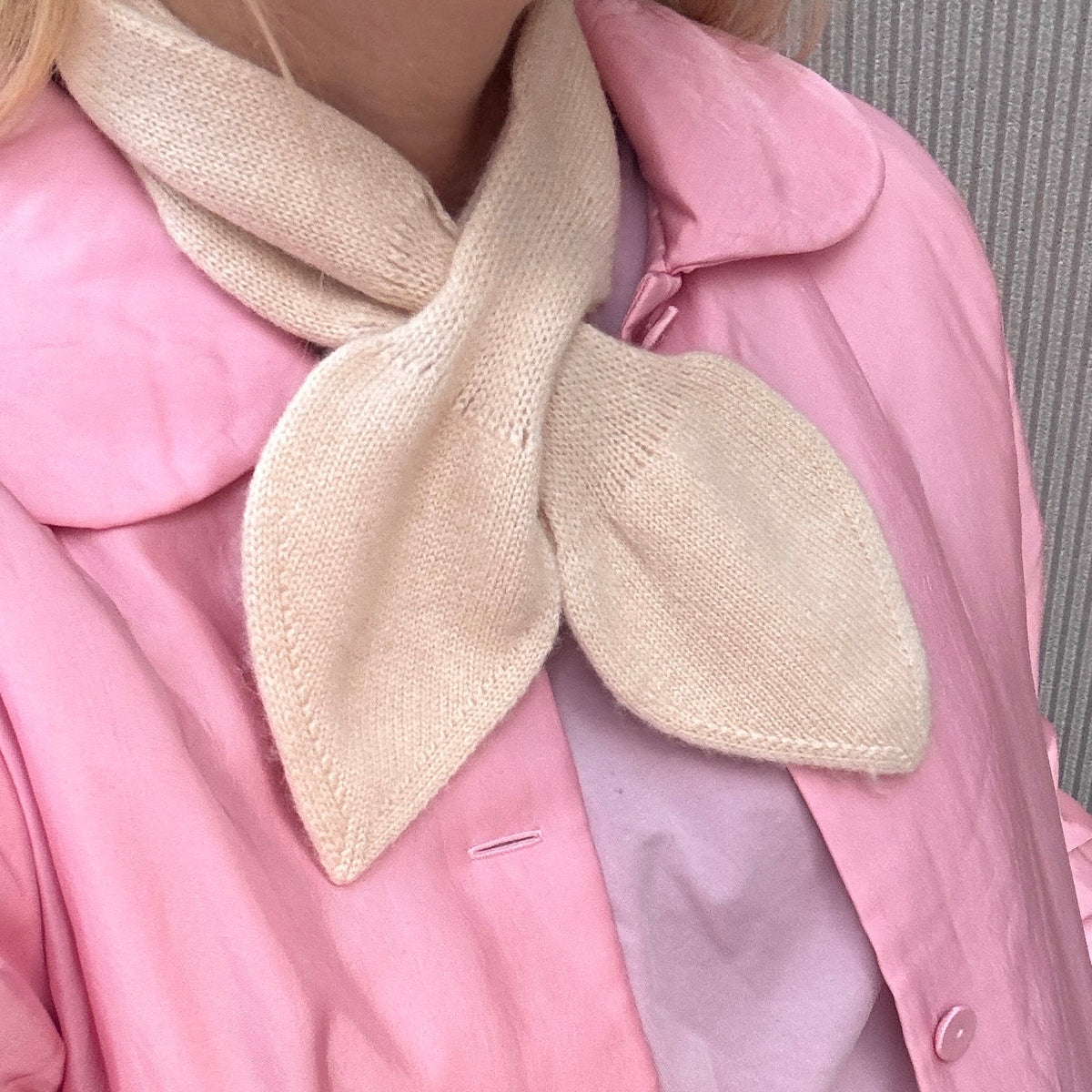 A close-up of a person wearing a light pink jacket over a pale pink top, with a large cream knit bow or scarf detail at the collar.