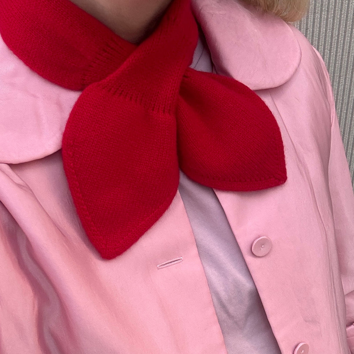 A close-up of a person wearing a light pink jacket with a large red bow tie detail, layered over a pale pink top.